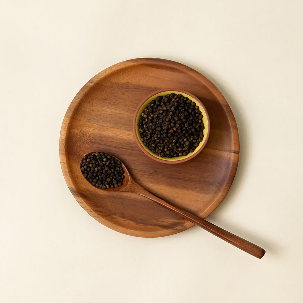 A small dish of whole black peppercorns is on top of a wood dinner plate, next to a wooden spoon which is partially covered by the whole black peppercorns as well.