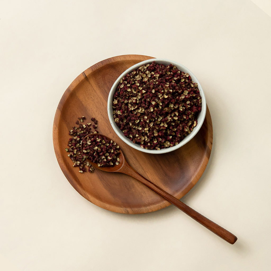 A small dish filled with Soeos Sichuan Peppercorns on top of a wood plate, next to a wooden spoon partially covered by the peppercorns.