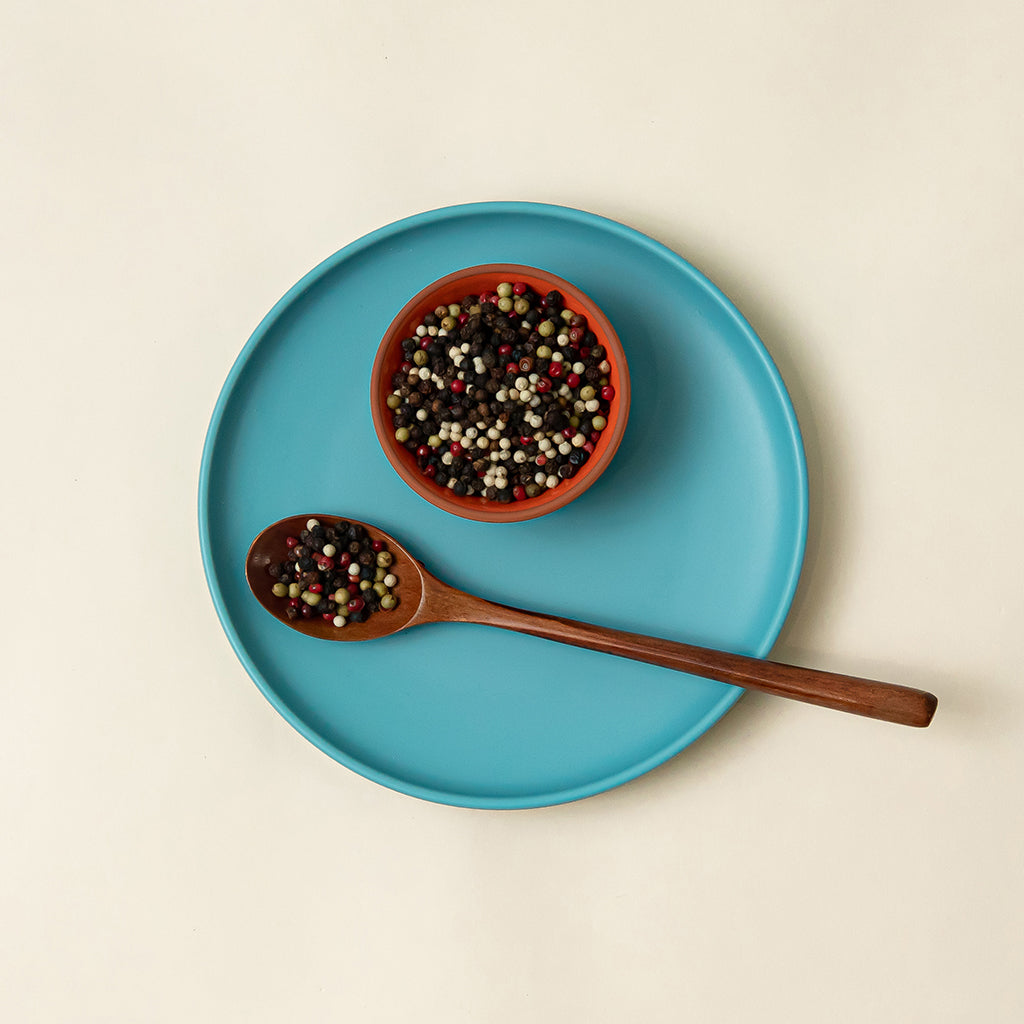On a blue plate, there are a wooden spoon and a wood dish which are full of rainbow peppercorns mix.