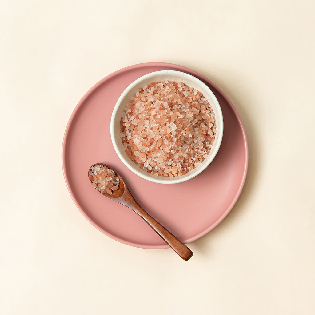 Soeos Pink Himalayan Salt is on top of a pink plate, next to a wooden spoon which is partially covered by the Pink Himalayan Salt.